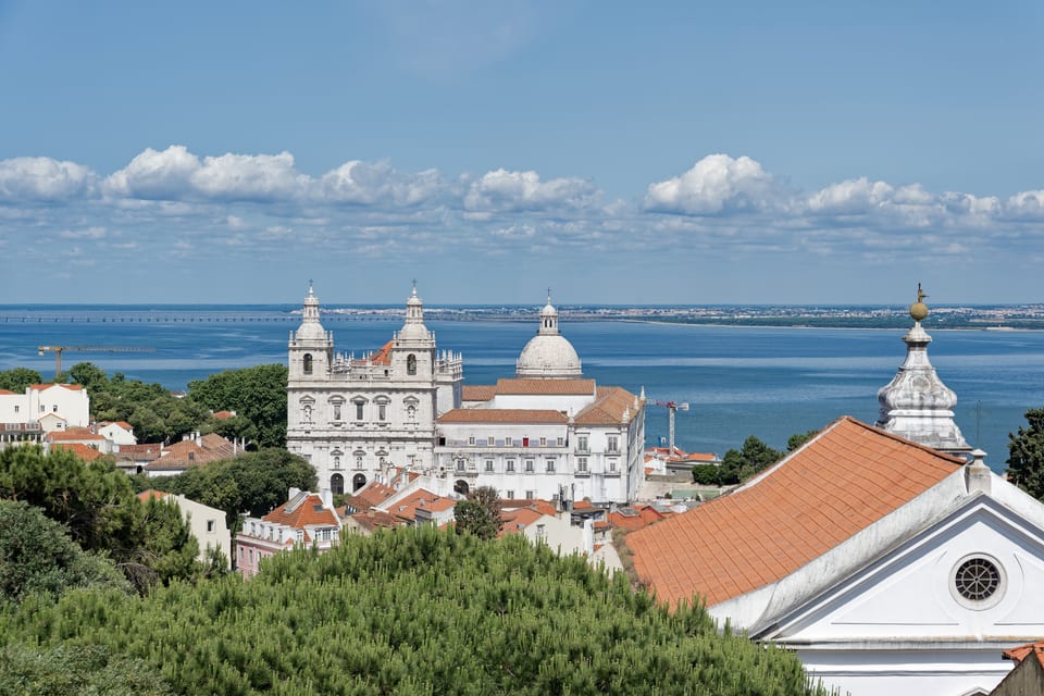 Lisbon seen from Sao Jorge Castle: terracotta rooftops falling away towards the Tagus estuary, with the twin white towers of the Sao Vicente de Fora monastery and the dome of the National Pantheon in the middle distance